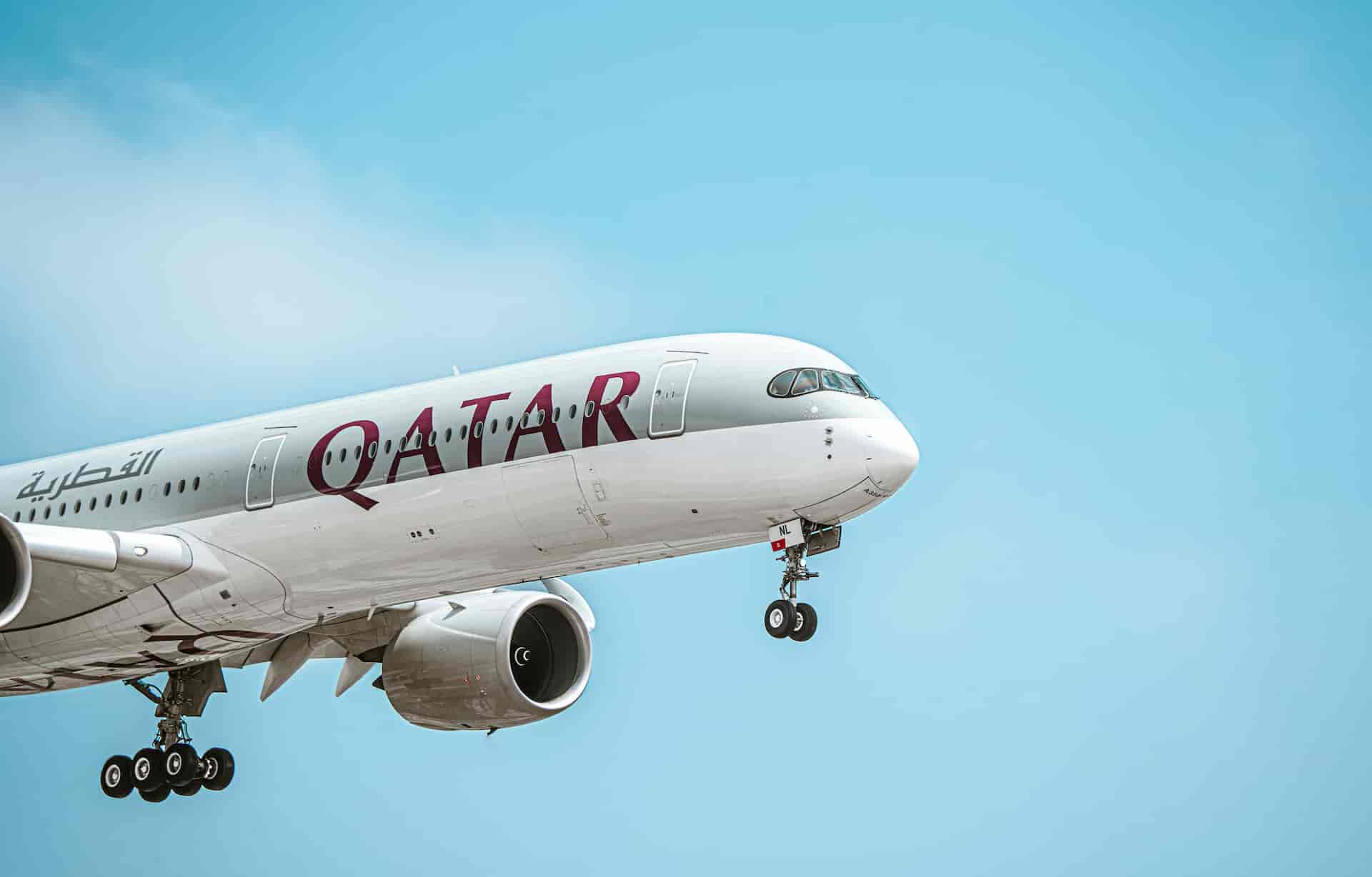 Qatar Airways wide-body airplane in flight with landing gear extended against a clear blue sky.