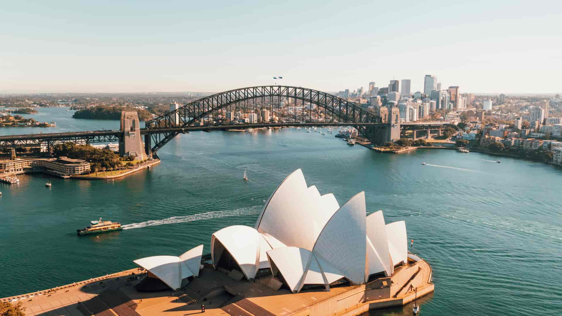 Aerial view of Sydney Harbour featuring the Sydney Opera House in the foreground, the Harbour Bridge spanning the water behind it, boats moving across the harbor, and the city skyline in the background under a clear sky.