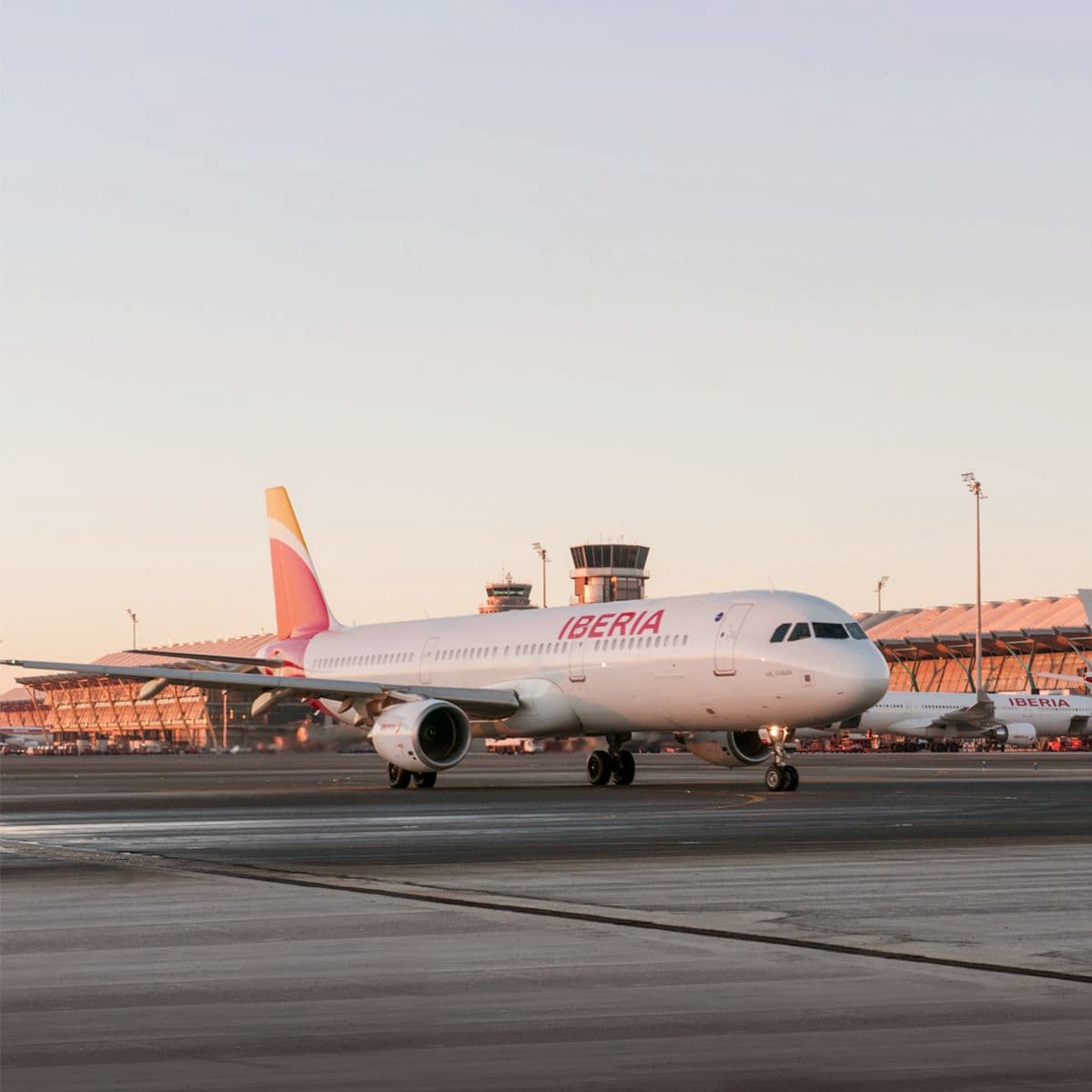 Iberia passenger airplane on a runway at an airport during sunset, with terminal buildings in the background.