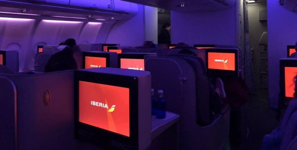 Business class cabin of an Iberia airplane with purple ambient lighting and individual screens displaying the Iberia logo.