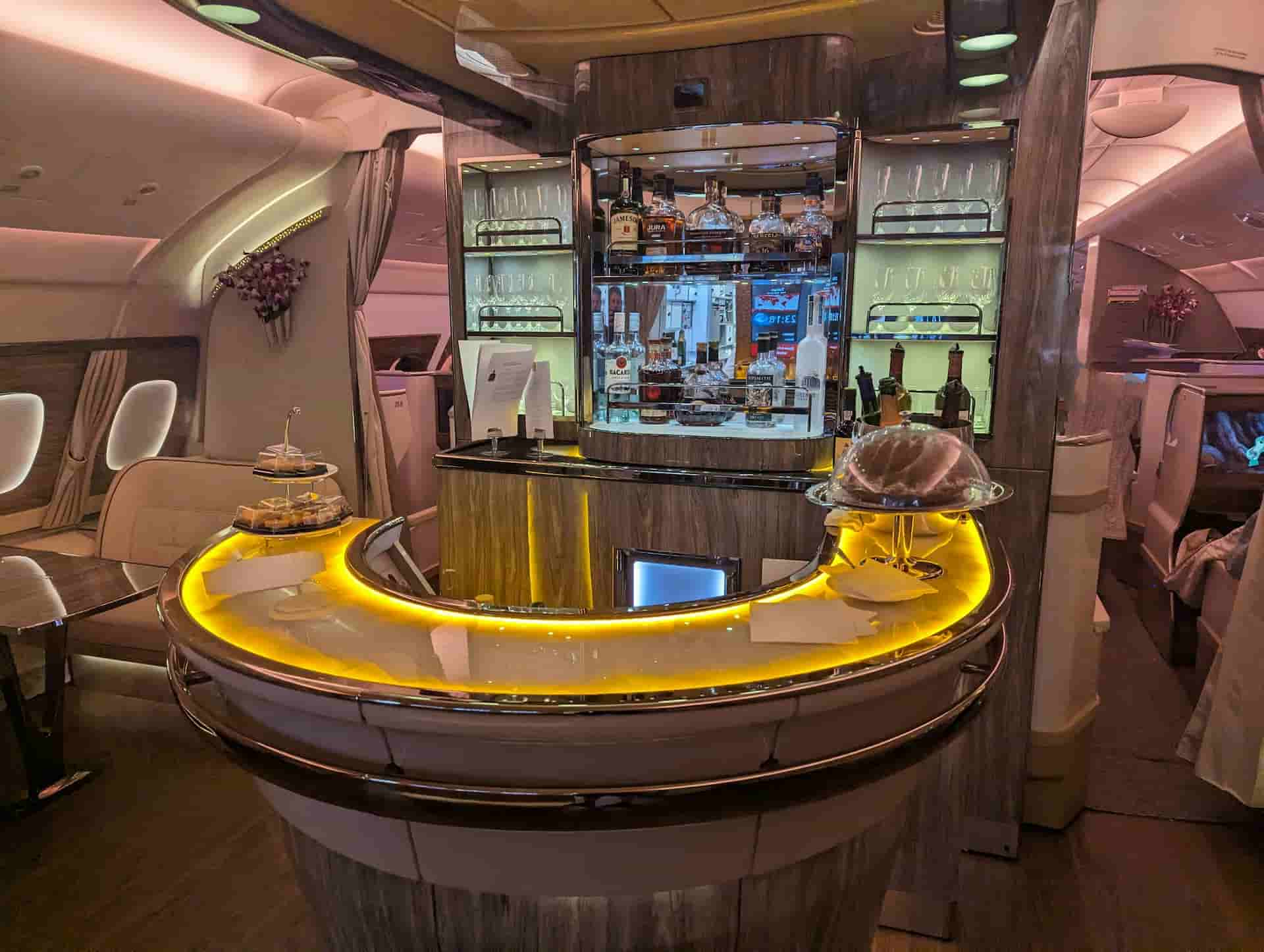 Luxury airplane onboard bar with curved illuminated counter, premium liquor display, and elegant seating area in a first-class cabin